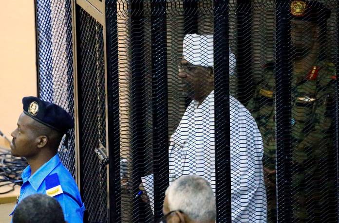 Sudan's former president Omar Hassan al-Bashir stands guarded inside a cage at the courthouse where he is facing corruption charges, in Khartoum