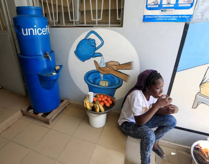 A girl eats as she sits next to a handwashing station, while she sells fruits at the entrance of a medical center in Pikine, on the outskirts of Dakar A girl eats as she sits next to a handwashing station, while she sells fruits at the entrance of a medical center in Pikine, on the outskirts of Dakar